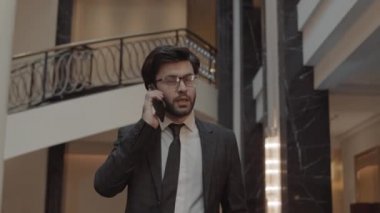 Koyu renk saçlı, gözlüklü, resmi kıyafet giyen, cep telefonuyla konuşan, otel lobisinde yürüyen genç bir işadamının belini dikizlemek.