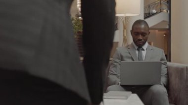 Orta uzunlukta, gri takım elbiseli, dizüstü bilgisayar kullanan ve otel lobisinde kendisine eşlik eden tanınmayan biriyle konuşan Afrikalı genç bir işadamı.