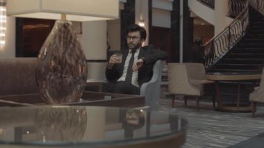 Koyu renk saçlı, gözlüklü ve özel dikim takım elbiseli genç bir işadamının izini sürüyorum. Saate bakıyorum, kayalardan içiyorum, koltukta oturuyorum, modern otel lobisinde bekliyorum.