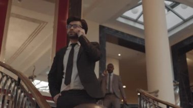 Düşük açılı başarılı CEO, özel yapım takım elbise ve gözlük takıyor, akıllı telefonla konuşuyor, arka planda merdivenlerden inen meslektaşı.