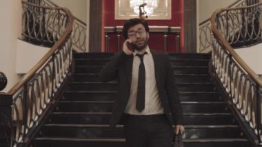 Koyu renk saçlı erkek girişimcinin resmi takım elbise ve gözlük takıp cep telefonuyla konuşurken, merdivenlerden inerken ve muhteşem otel odasında