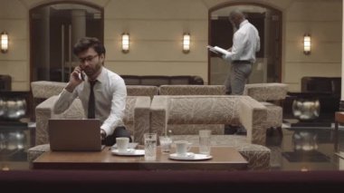 Orta uzunlukta başarılı genç bir CEO restorandaki koltukta dizüstü bilgisayarla oturuyor, cep telefonuyla konuşuyor, iş arkadaşı arka planda yürüyor.