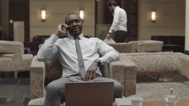 Orta uzunlukta bir Afrikalı girişimci restoran koltuğunda dizüstü bilgisayarla oturuyor, cep telefonuyla konuşuyor, iş arkadaşı arka planda yürüyor.