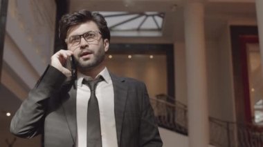 Düşük açılı, koyu saçlı, kendine güvenen ofis çalışanı cep telefonuyla konuşuyor, yüksek tavanlı şık bir yerde duruyor.