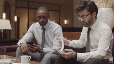 Orta uzunlukta iki genç erkek iş ortağının kontrat imzalaması ve el sıkışması, şık bir restoranda koltukta oturup konuşması.