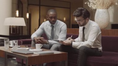 Orta uzunlukta iki genç erkek iş ortağı kontrat imzalıyor ve el sıkışıyor, şık bir restoranda kanepede oturuyor.