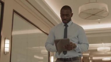 Beyaz gömlek ve kravat giyen, tablet bilgisayar kullanan, iş merkezinin koridorunda yürüyen, düşük açılı genç Afrikalı erkek yöneticinin orta açı takibi.