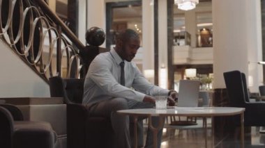 Orta uzunlukta bir Afrikalı işadamı resmi kıyafetler giyiyor, dizüstü bilgisayarda yazıyor, içme suyu içiyor, iş merkezinin lobisindeki koltukta oturuyor.