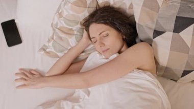 Koyu saçlı, beyaz bir kadının yatak odasında uyuduğunu, el pençe divan durduğunu, cep telefonunu yatakta kapatmaya çalıştığını, sonra kaşlarını çattığını ve tekrar uykuya daldığını.