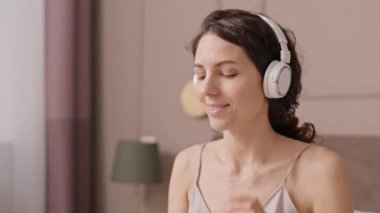 Göğsü kabarık, beyaz bir kadın kulaklık takıyor, akıllı telefon kullanıyor, müzik dinliyor ve odasında oturup eğleniyor.