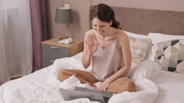 Yüksek açılı beyaz kadın yatakta oturuyor, bilgisayarında video görüşmesi yapıyor, gülümsüyor ve el sallıyor,