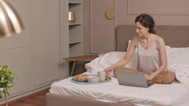 大大的棕色眼睛的白人女人坐在床上 使用笔记本电脑 喝咖啡 早餐时把杯子放在盘子里 — 图库视频影像