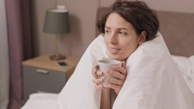 Yüksek açılı, kahverengi saçlı beyaz kadın battaniyeye sarılmış, sıcak kahvenin keyfini çıkarıyor ve ısınıyor.