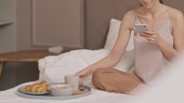 Pijama giyen, yatakta oturan, akıllı telefon kullanan, kahvaltıda tepsiden kupa alan, kahve içen güzel beyaz bir kadın.