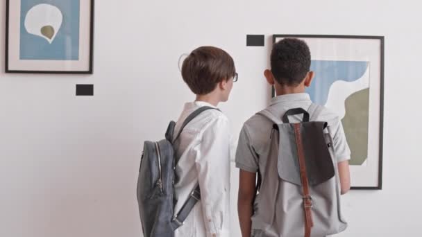 バックパックを着た2人の多民族少年の後ろ姿 美術館での絵画の観察 — ストック動画
