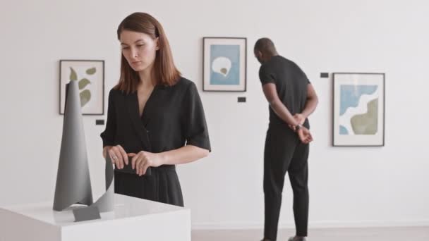 被锁定的白人年轻女子用智能手机在画廊里拍照 然后离去 男人在后台观看绘画 — 图库视频影像