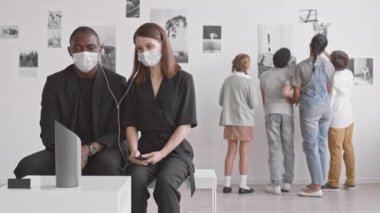 Orta uzunlukta, siyah giysiler ve tıbbi maskeler takan, modern sanat galerisinde oturan, kulaklıkla ses rehberini dinleyen, arka planda duran çocuklar.