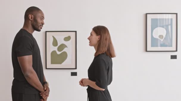 穿着黑色衣服的高个子非洲男人和白人女人站在艺术画廊边 讨论墙上的现代绘画 — 图库视频影像
