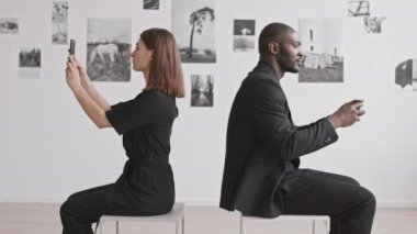 Kafkasyalı kadın ve Afrikalı erkeğin fotoğraf sergisindeki taburelerin üzerinde oturup akıllı telefonlarla fotoğraf çektikleri ve birbirlerine resimlerini gösterdikleri orta boy bir manzara.