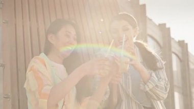 İki neşeli Asyalı ve Kafkasyalı kız arkadaşın sıcak yaz gününde dışarıda limonata içip sohbet ederken orta boy bir fotoğrafı.