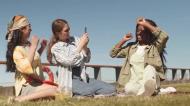 Neşeli Afrikalı Afrikalı Amerikalı kadın, çok ırklı bayan arkadaşları sahilde çimlerin üzerinde otururken komik dans hareketleri sergiliyor.