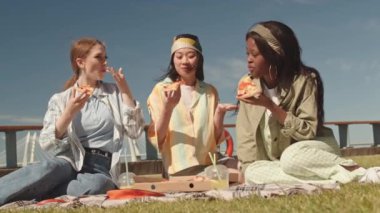 Beyaz, Asyalı ve Afrikalı Amerikalı genç kadınlar parktaki çimlerde ya da açık mavi gökyüzünün altında birlikte oturmuş sohbet ediyor ve pizza ve limonatanın tadını çıkarıyorlar.