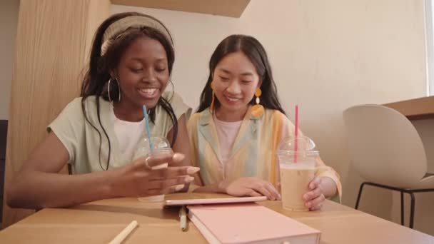 デジタルタブレットを見て会話をしている2人の若い多民族女性のウエストアップショット プラスチックカップのアイスコーヒーとカフェテーブルに座って — ストック動画