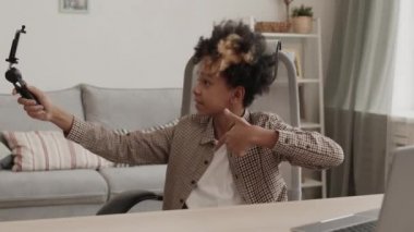 Neşeli, Afro-Amerikan ergen bir çocuk evinde oturmuş elinde akıllı bir telefonla video blogu için kendini videoya çekiyor.