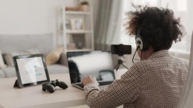 Genç Afrikalı çocuğun evdeki masasında oturup taşınabilir bilgisayarda online oyun oynaması, sonra sandalyeye dönüp kameraya gülümsemesi.