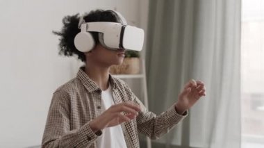 Orta uzunlukta günlük kıyafetler giyen ve VR kulaklık takan, odada duran, başını çeviren ve el sallayan, sonra gözlüğünü çıkaran, heyecanlı görünen genç bir çocuk.