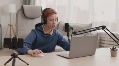 Mavi kapüşonlu ve kulaklıklı kızıl saçlı çocuk, odasında oturmuş, dizüstü bilgisayarla bilgisayar oyunu oynuyor, blog için akıllı telefondan video çekiyor.