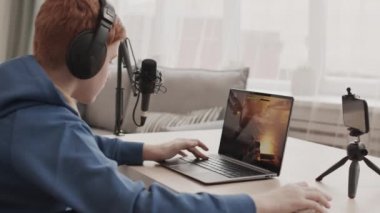 Kulaklık takan kızıl saçlı bir çocuğun yan görünüşü odada masada oturması, taşınabilir bilgisayarda atış oyunu oynaması, akıllı telefon kamerasıyla kayıt yapması.
