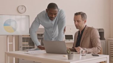 Orta yaşlı, beyaz bir adam ofisinde oturuyor, erkek Afrikalı meslektaşı yanında duruyor, konuşuyor ve taşınabilir bilgisayar ekranını işaret ediyor.