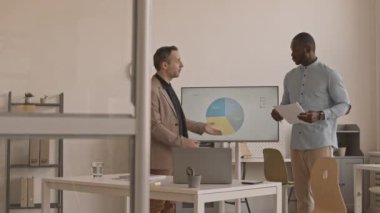 Çok ırklı iki erkek meslektaşın resmi olarak gündüz ofiste dikilip dizüstü bilgisayarla video konferansı yapması, sunum yapması, konuşması...