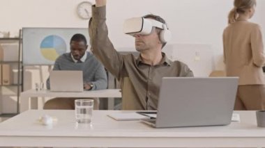 Ofiste masasında oturan, sanal gerçeklik kulaklığı takan, elini hareket ettiren beyaz bir adamın yanından geçen...