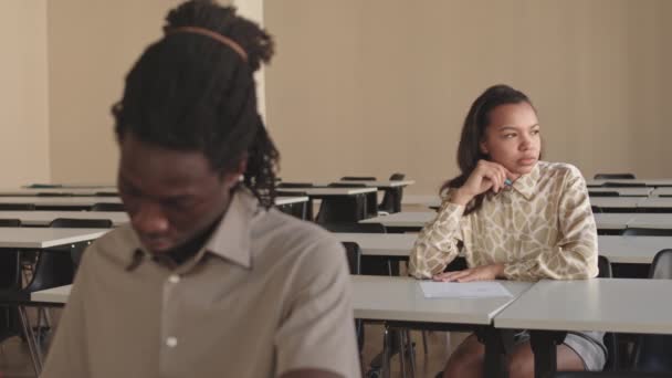 女子大生の中長期教室の学校の机に座って 考えて ぼやけたアフリカの同級生の背景に試験に書きます — ストック動画