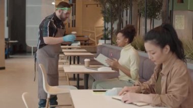 Kafkas garson önlük, eldiven ve yüz kalkanı ile genç bayan melez ve Asyalı müşterilere sıcak içecek ve kağıt bardaklar servis ediyor.