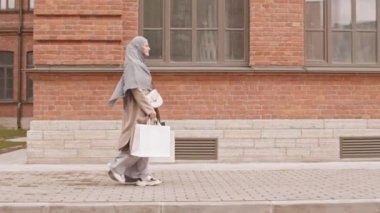 Ağır çekimde yan görüntü takibi ya da tesettürlü modern Arap kadın alışveriş torbaları taşıyarak sokakta yürüyor.
