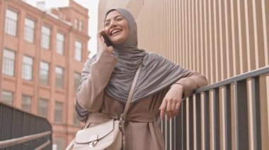 Şehir ortamında telefon görüşmeleri sırasında tesettürlü, mutlu, genç bir Arap kadının gülüşünün orta yavaş görüntüsü.