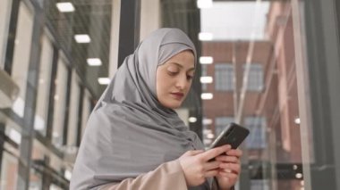 Modern ofis binasının içinde duran, tesettürlü, çekici, genç bir Arap kadının yavaş çekimleri ile düşük açılı orta açılı, yakın çekim.