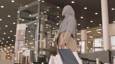 Modern alışveriş merkezinin önünde duran tesettürlü ve alışveriş çantalı çağdaş Arap kadının düşük açılı yavaş çekimi.