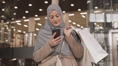 Orta kavisli mutlu, tesettürlü ve alışveriş çantalı genç bir Arap kadının kapalı bir alışveriş merkezinde kapalı bir şekilde dururken çektiği fotoğraf.