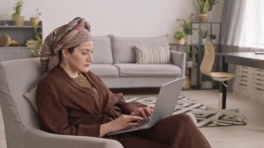 Modern türbanlı gülümseyen Müslüman kadının evdeki sandalyede oturarak dizüstü bilgisayarda çalışmasının orta derece yavaşlaması.