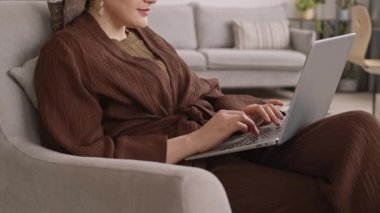 Sarıklı ve büyük küpeli gülümseyen Müslüman kadının rahat dairesinde dizüstü bilgisayarla vakit geçirirken yavaş çekimleri.