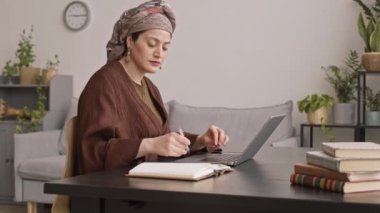 Yan görüntüde, türban başlıklı modern Müslüman bir kadının laptopta çalışırken çekilmiş resmi var. Kitaptan notlar alıyor.