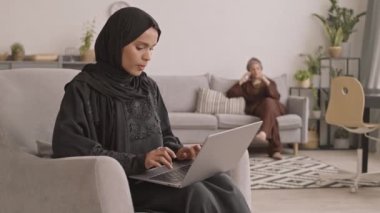 Siyah elbiseli, tesettürlü, tesettürlü, mutlu, genç bir kadının laptopla çalışırken, kız arkadaşının telefonda arka planda koltukta oturup konuşmasının yavaş çekimleri.