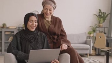 Evde dizüstü bilgisayar başında komik video ya da film izlerken türbanlı ve tesettürlü iki neşeli Arap kadının yavaş çekimleri.