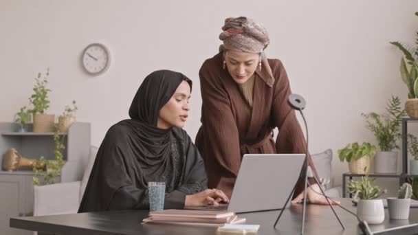 明るい居心地の良いアパートでラップトップで作業しながら 伝統的な服やスカーフチャットで2人の魅力的なアラビア人女性のスローショット — ストック動画