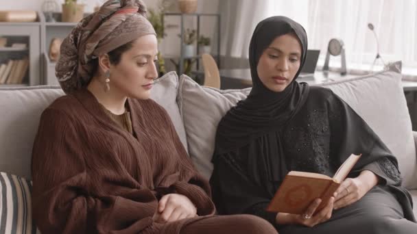 中低速ショットの二人の若いですムスリム女性読みますコーラン座っています上のソファに家 — ストック動画