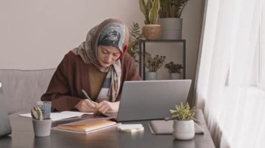 Türbanlı genç Müslüman bir kadının, modern apartmandaki dizüstü bilgisayarın önünde masa başında evrak işleriyle uğraşması.